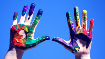 Mãos no ar com tintas de várias cores com o céu azul por trás